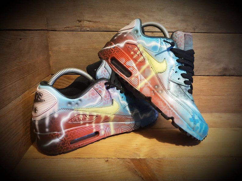 May include: A pair of custom-painted Nike Air Max 90 sneakers. One shoe features a blue, yellow, and red lightning bolt design, while the other shoe features a red, yellow, and blue lightning bolt design.