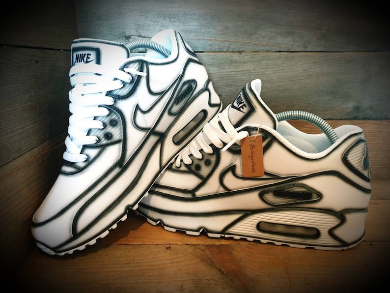 May include: White Nike Air Max 90 sneakers with black outlines. The shoes have white laces and a white swoosh logo. The tag on the shoe reads "Reimagined".