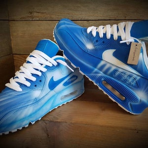 May include: A pair of blue and white custom-painted Nike Air Max 90 sneakers with white laces. The shoes have a white swoosh logo and a blue Air Max logo on the side.