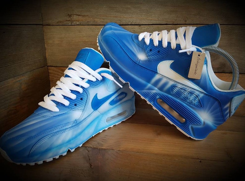 May include: A pair of blue and white custom-painted Nike Air Max 90 sneakers with white laces. The shoes have a white swoosh logo and a blue Air Max logo on the side.