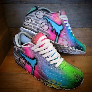May include: A pair of custom-painted Nike Air Max 90 trainers. One shoe features a grey and white base with black and white cartoon faces, while the other shoe has a pink, blue, and green base with a galaxy-like design.
