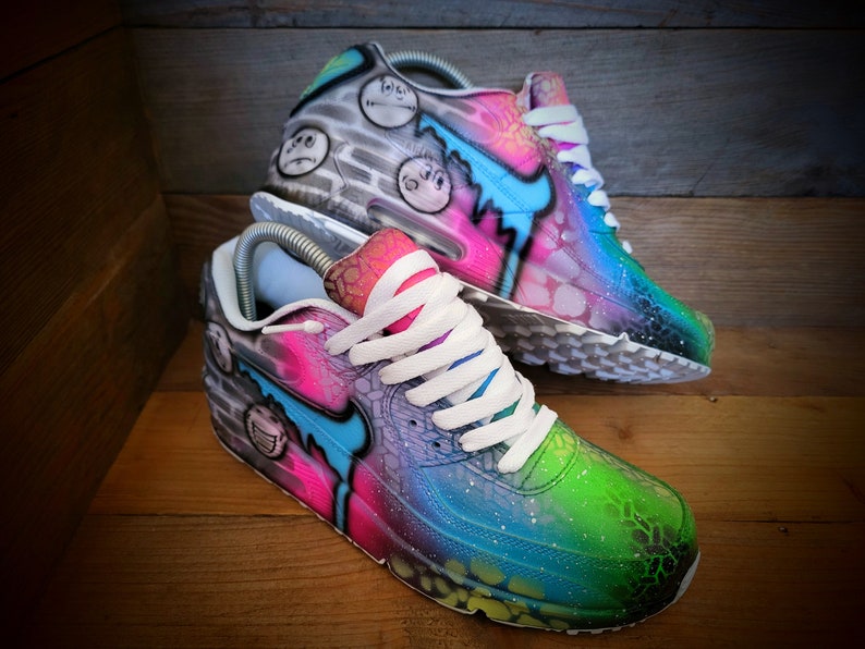 May include: A pair of custom-painted Nike Air Max 90 trainers. One shoe features a grey and white base with black and white cartoon faces, while the other shoe has a pink, blue, and green base with a galaxy-like design.