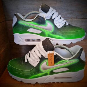 May include: A pair of white and green custom-painted Nike Air Max 90 sneakers with a black swoosh and a lightning bolt design. The shoes have a white midsole and a black outsole.