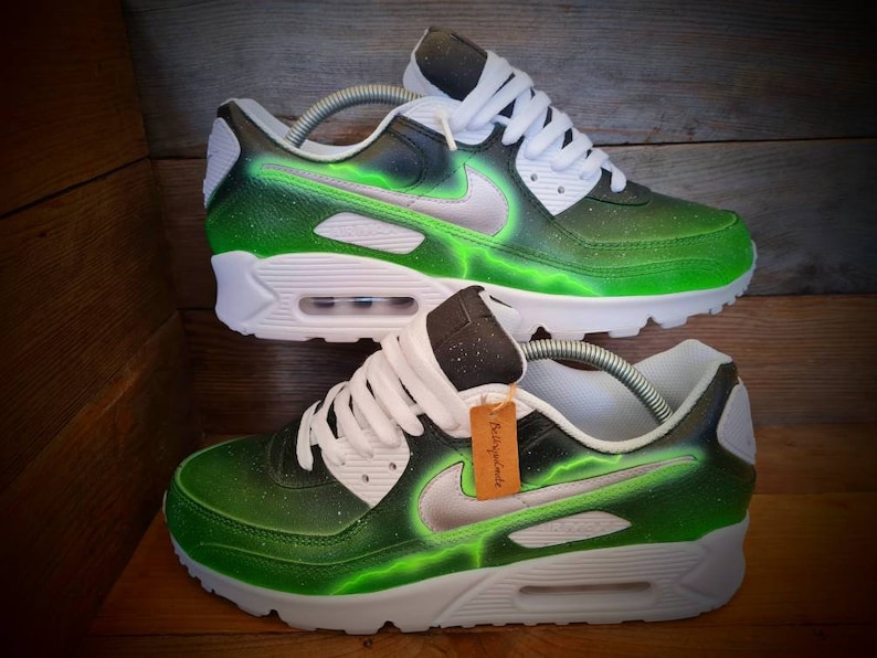 May include: A pair of white and green custom-painted Nike Air Max 90 sneakers with a black swoosh and a lightning bolt design. The shoes have a white midsole and a black outsole.