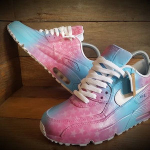 May include: A pair of white sneakers with a pink and blue ombre design. The sneakers have a star pattern on the pink side and a white swoosh logo on the blue side.