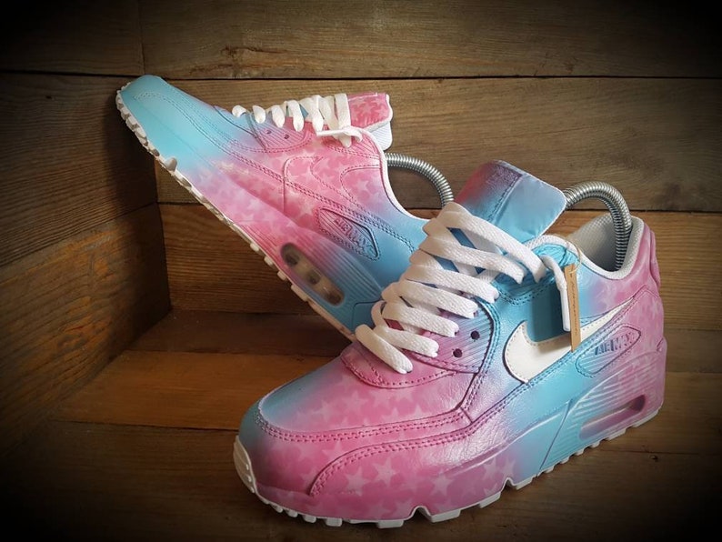 May include: A pair of white sneakers with a pink and blue ombre design. The sneakers have a star pattern on the pink side and a white swoosh logo on the blue side.