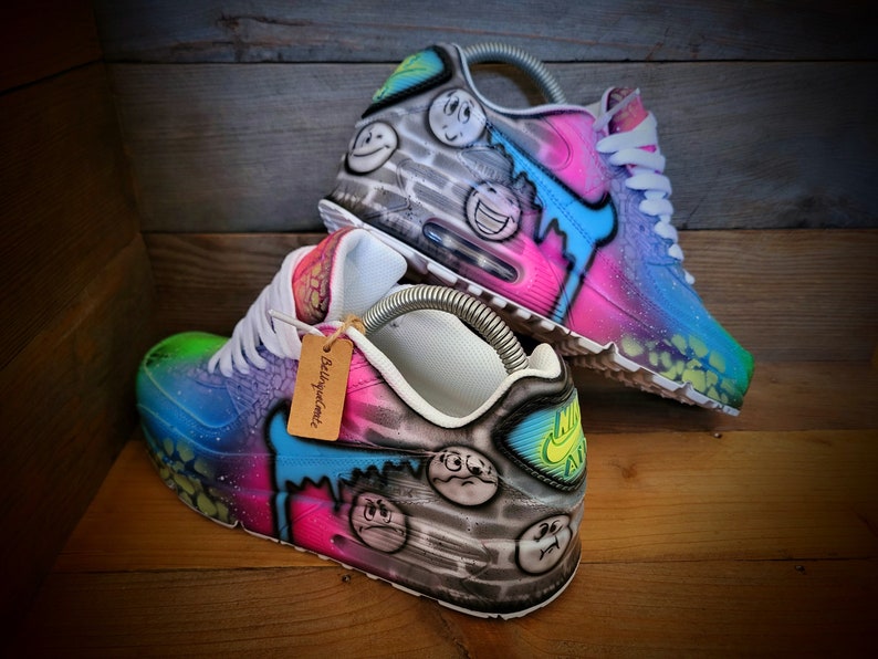 May include: A pair of custom-painted Nike Air Max 90 trainers with a galaxy-inspired design. The shoes feature a black and white brick pattern with smiley faces, a blue and green gradient, and pink accents. The tag on the shoe reads "Bellejournee Art".