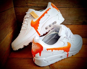 Könnte beinhalten: Weiße Lederschuhe mit orangefarbenen Tropf-Farbdetails. Die Schuhe haben ein weißes Swoosh-Logo und den Text "Air" an der Seite.