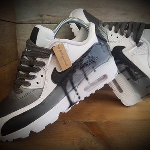 May include: A pair of white and gray sneakers with black accents. The sneakers have a black swoosh logo and the text "Air Max" on the side. The sneakers are custom painted with a black and white abstract design.