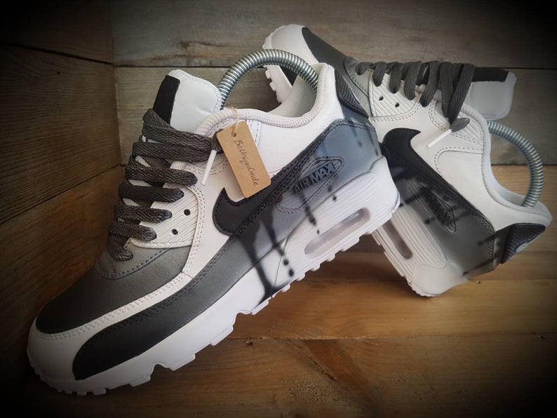 May include: A pair of white and gray sneakers with black accents. The sneakers have a black swoosh logo and the text "Air Max" on the side. The sneakers are custom painted with a black and white abstract design.