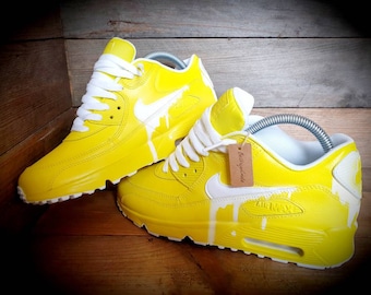 Könnte beinhalten: Ein Paar gelbe Sneaker mit weißen Akzenten. Die Sneaker haben ein weißes Swoosh-Logo und weiße Farbtropfen. Der Text "Air Max" ist an der Seite des Schuhs sichtbar. Das Etikett am Schuh lautet "Betterhandmade".