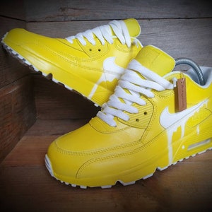 May include: A pair of yellow leather sneakers with white laces and a white swoosh logo. The sneakers have a white paint drip design on the side.