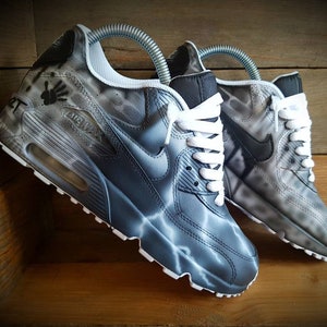 May include: A pair of custom-painted white and gray Nike Air Max 90 sneakers with black accents. The sneakers feature a unique lightning bolt design and a handprint on the side.