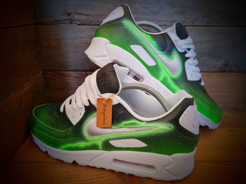 May include: Green and white custom-painted Nike Air Max 90 sneakers with a lightning bolt design. The shoes have a black swoosh and a white midsole.