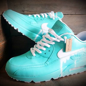 May include: A pair of turquoise blue Nike Air Max 90 sneakers with white laces, a white swoosh logo, and white paint splatters on the side. The sneakers are on a wooden surface.