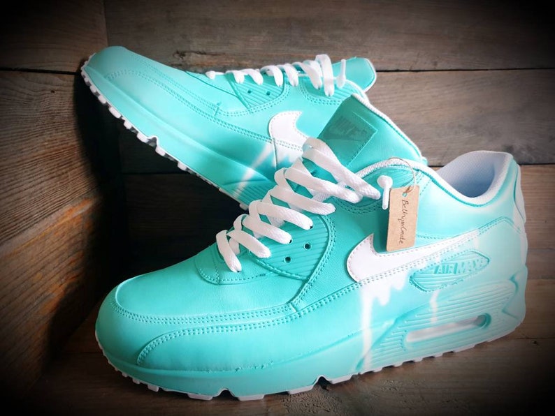 May include: A pair of turquoise blue Nike Air Max 90 sneakers with white laces, a white swoosh logo, and white paint splatters on the side. The sneakers are on a wooden surface.