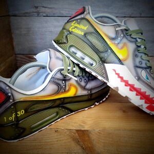 May include: Custom-painted green, gray, and yellow Nike Air Max 90 sneakers with a red and white shark tooth design on the sole. The sneakers are labeled "Limited Edition" and "1 of 30".