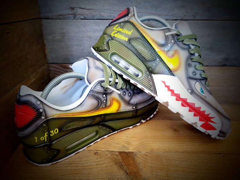 May include: Custom-painted green, gray, and yellow Nike Air Max 90 sneakers with a red and white shark tooth design on the sole. The sneakers are labeled "Limited Edition" and "1 of 30".