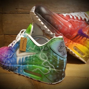 May include: A pair of custom-painted Nike Air Max 90 sneakers. One shoe is painted with a rainbow color scheme, while the other is painted with a green, blue, and purple color scheme. Both shoes have a white midsole and a black outsole.