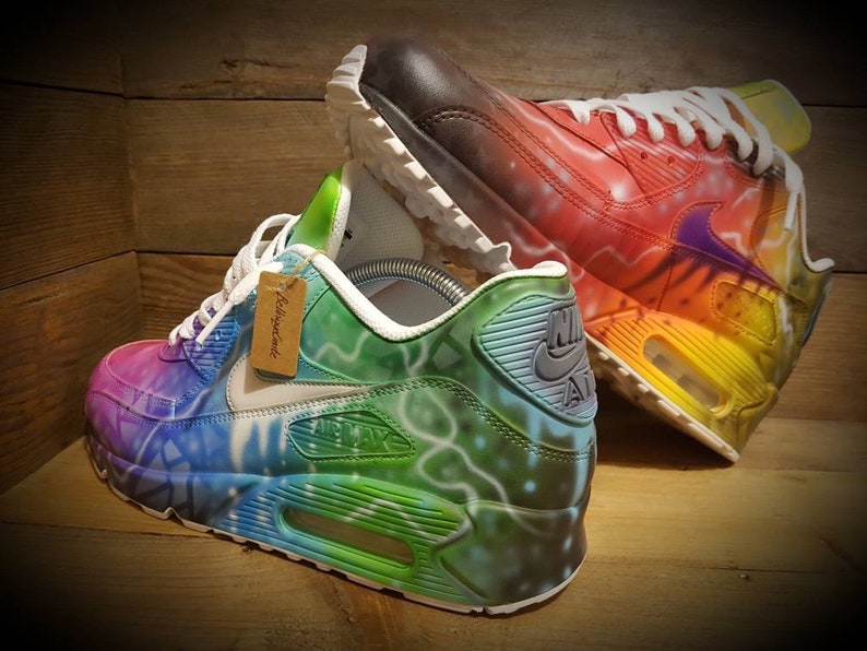May include: A pair of custom-painted Nike Air Max 90 sneakers. One shoe is painted with a rainbow color scheme, while the other is painted with a green, blue, and purple color scheme. Both shoes have a white midsole and a black outsole.