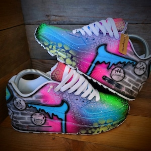 May include: A pair of custom-painted white trainers with a blue, green, pink, and black galaxy-inspired design. The trainers feature a variety of smiley faces and other graffiti-style artwork.