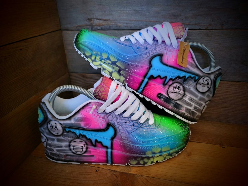 May include: A pair of custom-painted white trainers with a blue, green, pink, and black galaxy-inspired design. The trainers feature a variety of smiley faces and other graffiti-style artwork.