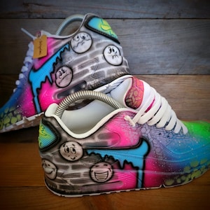 May include: A pair of custom-painted white trainers with a colourful design featuring smiley faces, drips, and a brick wall pattern. The trainers are a vibrant mix of pink, blue, green, and yellow.