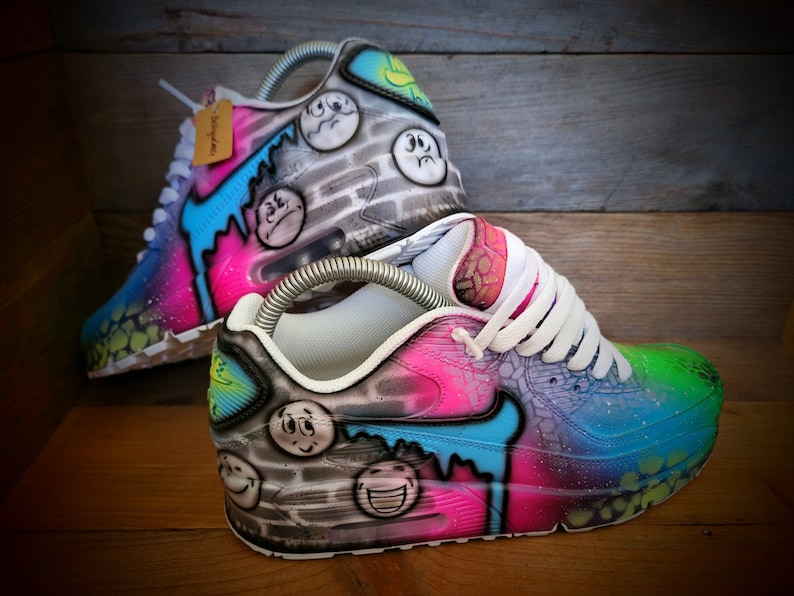 May include: A pair of custom-painted white trainers with a colourful design featuring smiley faces, drips, and a brick wall pattern. The trainers are a vibrant mix of pink, blue, green, and yellow.