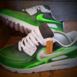 May include: Custom-painted green and black Nike Air Max 90 sneakers with a silver swoosh and a green lightning bolt design. The shoes have white laces and a white midsole.