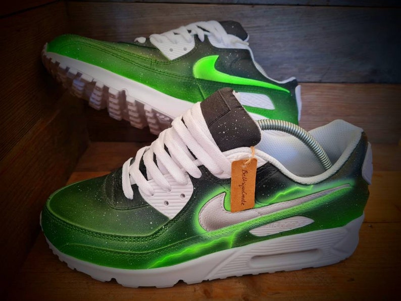 May include: Custom-painted green and black Nike Air Max 90 sneakers with a silver swoosh and a green lightning bolt design. The shoes have white laces and a white midsole.