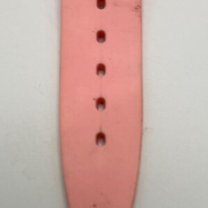 Puede incluir: Primer plano de una correa de reloj rosa claro con varios agujeros para ajustar. La correa muestra signos de desgaste, con algo de decoloraci&oacute;n y peque&ntilde;os da&ntilde;os en el extremo. La correa est&aacute; hecha de un material flexible.