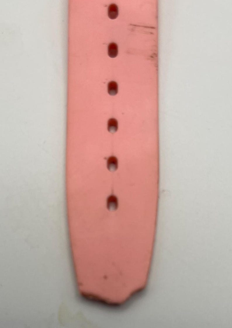 Puede incluir: Primer plano de una correa de reloj rosa claro con varios agujeros para ajustar. La correa muestra signos de desgaste, con algo de decoloraci&oacute;n y peque&ntilde;os da&ntilde;os en el extremo. La correa est&aacute; hecha de un material flexible.