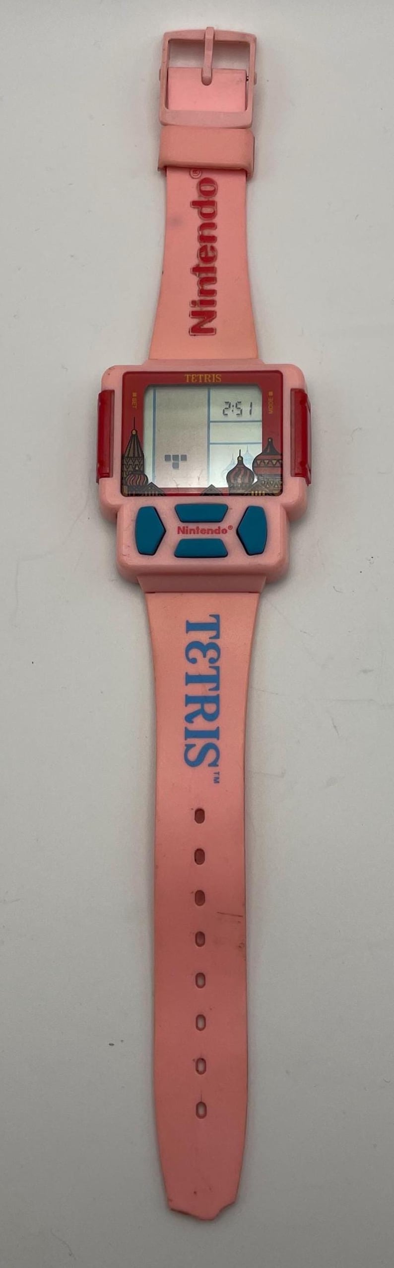 Puede incluir: Reloj Nintendo Tetris rosa con una pantalla digital que muestra la hora. El reloj presenta una correa rosa con la palabra "TETRIS" y el logotipo de Nintendo. La esfera del reloj tiene botones azules y un borde rojo.