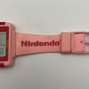 Puede incluir: Un reloj Nintendo Tetris rosa con una correa y hebilla rosas a juego. La esfera del reloj muestra el juego Tetris y el logotipo de Nintendo est&aacute; impreso en la correa.