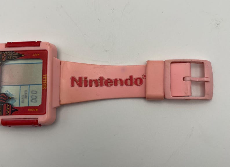 Puede incluir: Un reloj Nintendo Tetris rosa con una correa y hebilla rosas a juego. La esfera del reloj muestra el juego Tetris y el logotipo de Nintendo est&aacute; impreso en la correa.