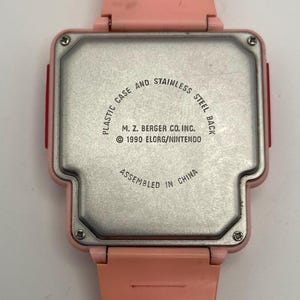 Puede incluir: Un reloj rosa vintage con parte trasera de acero inoxidable. La caja del reloj es de pl&aacute;stico. La parte trasera del reloj tiene el texto "PLASTIC CASE AND STAINLESS STEEL BACK", "M. Z. BERGER CO. INC. &copy; 1990 ELORG/NINTENDO" y "ASSEMBLED IN CHINA".