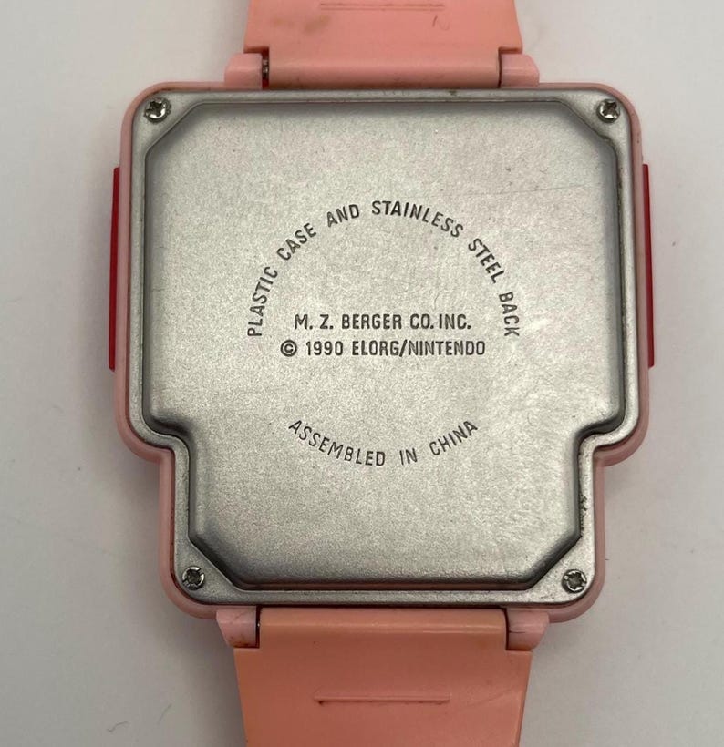 Puede incluir: Un reloj rosa vintage con parte trasera de acero inoxidable. La caja del reloj es de pl&aacute;stico. La parte trasera del reloj tiene el texto "PLASTIC CASE AND STAINLESS STEEL BACK", "M. Z. BERGER CO. INC. &copy; 1990 ELORG/NINTENDO" y "ASSEMBLED IN CHINA".