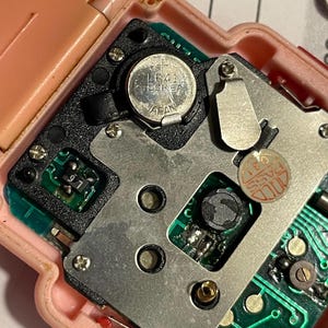 Puede incluir: Primer plano de los componentes internos de un peque&ntilde;o dispositivo electr&oacute;nico. Una placa de metal plateado est&aacute; asegurada con tornillos, y una bater&iacute;a circular LR41 Toshiba es visible. El dispositivo tiene una carcasa de pl&aacute;stico rosa y placas de circuito verdes.