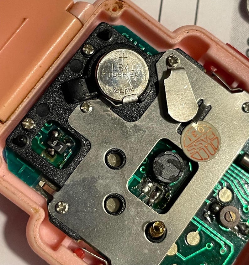 Puede incluir: Primer plano de los componentes internos de un peque&ntilde;o dispositivo electr&oacute;nico. Una placa de metal plateado est&aacute; asegurada con tornillos, y una bater&iacute;a circular LR41 Toshiba es visible. El dispositivo tiene una carcasa de pl&aacute;stico rosa y placas de circuito verdes.