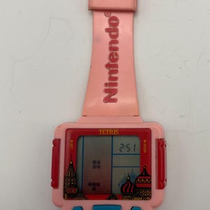 Puede incluir: Un reloj Nintendo Tetris rosa vintage. La esfera del reloj muestra el juego Tetris y la hora 2:51. La correa y la esfera son rosas con botones azules y el logotipo de Nintendo.