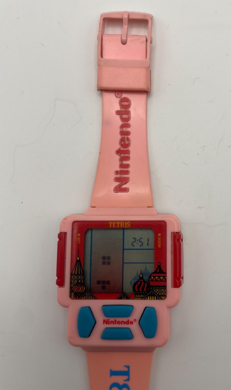 Puede incluir: Un reloj Nintendo Tetris rosa vintage. La esfera del reloj muestra el juego Tetris y la hora 2:51. La correa y la esfera son rosas con botones azules y el logotipo de Nintendo.