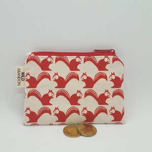 May include: A small, rectangular coin purse with a red zipper and a repeating pattern of red squirrels on a cream background. The tag on the side reads "WILD ABANDON". Two gold coins are in front of the purse.