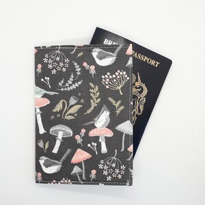 May include: A dark grey passport holder featuring a pattern of birds, mushrooms, and floral designs in pink, white, and beige. A British passport is partially visible behind the holder. The holder is made of fabric.