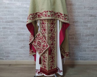 May include: A green liturgical vestment with red embroidered floral designs. The vestment has a long, flowing skirt and a cape-like top. A small, matching pouch hangs from the side.