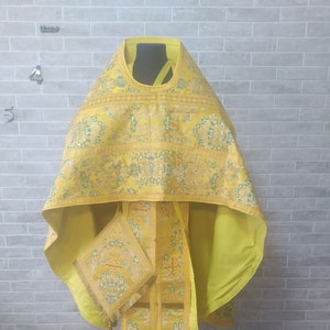 May include: A yellow liturgical vestment with gold embroidery and a matching stole. The vestment is displayed on a tripod stand against a brick wall.