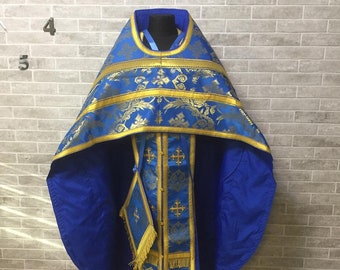 May include: A blue and gold liturgical vestment with a decorative pattern. The vestment is displayed on a stand against a brick wall.
