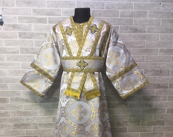 Puede incluir: Una túnica litúrgica blanca con bordados dorados y azules. La túnica tiene un patrón circular con cruces y se lleva puesta en un maniquí.