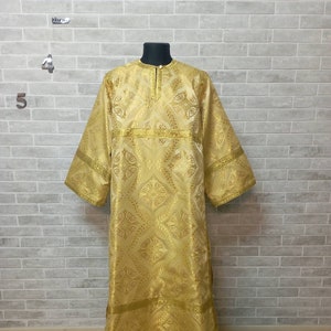 May include: A gold liturgical robe with a patterned design and gold trim. The robe is displayed on a stand against a brick wall.