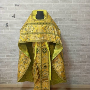 May include: A yellow and green liturgical vestment with intricate embroidery and gold accents. The vestment is displayed on a stand against a brick wall.