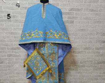 May include: A light blue liturgical vestment with gold embroidery. The vestment has a long, flowing design with a decorative collar and a matching stole. The stole has a small, square pouch attached to it.